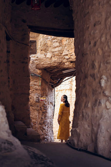 Back view of young female traveler in yellow dress standing in doorway of antique ruined stone house in old village Al Hamra in Oman — Stock Photo