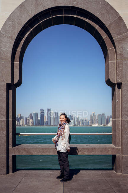 Vista lateral de la alegre turista asiática sonriendo ante la cámara mientras está parada sola junto a una valla arqueada en el paseo marítimo en el Museo de Arte Islámico contra West Bay y una maravillosa vista del centro de Doha - foto de stock