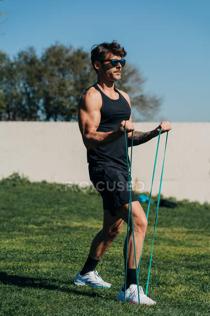 Strong male athlete in sportswear and sunglasses exercising with elastic band on lawn in sunlight — Stock Photo