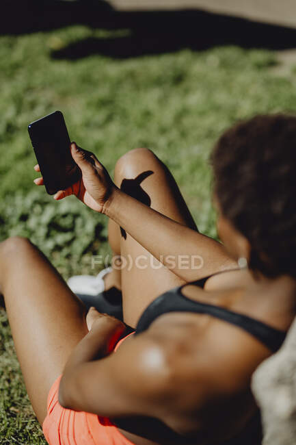 Неузнаваемая афроамериканка в спортивной одежде держит смартфон, сидя на траве в солнечный день — стоковое фото