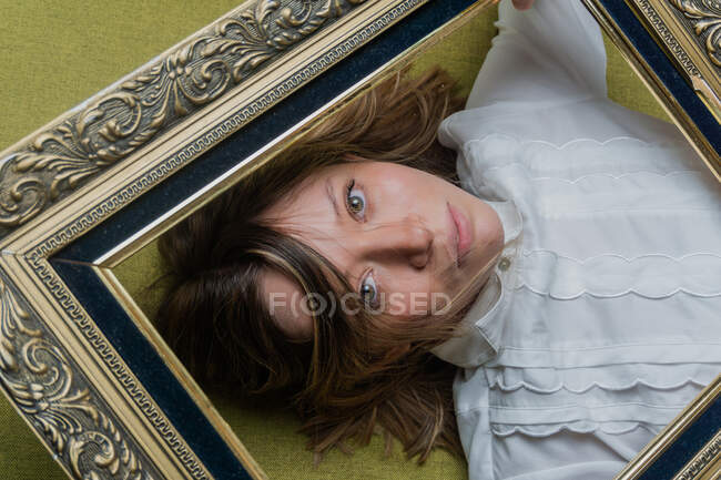 Vista dall'alto della giovane donna dai capelli lunghi con gli occhi verdi vestiti di camicetta bianca che guarda la fotocamera attraverso la cornice ornamentale — Foto stock