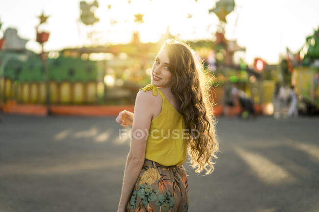 Задоволена жінка стоїть, дивлячись на камеру на ярмарку і насолоджуючись літніми вихідними під час заходу сонця — стокове фото