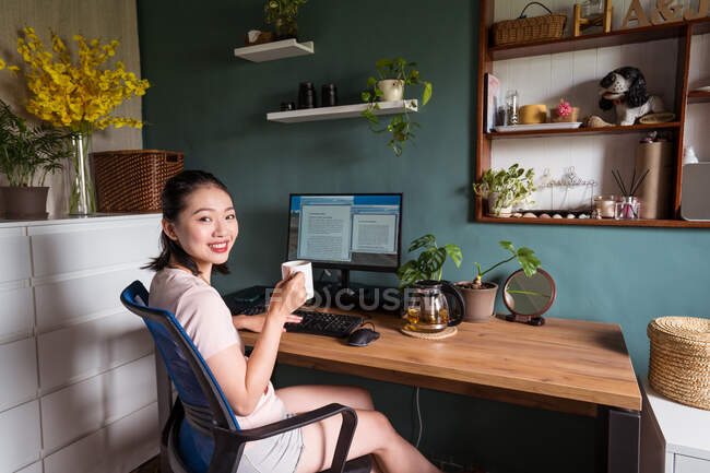 Vista lateral da feliz asiática freelancer leitura de documentos no monitor do computador enquanto sentado olhando para a câmera na mesa e bebendo chá durante o trabalho remoto — Fotografia de Stock