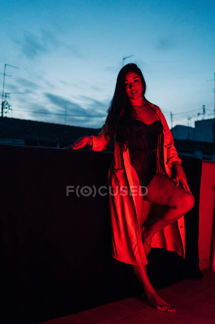 Delighted young Hispanic ethnic female in lingerie looking at camera while resting on terrace under red neon light at night — Stock Photo
