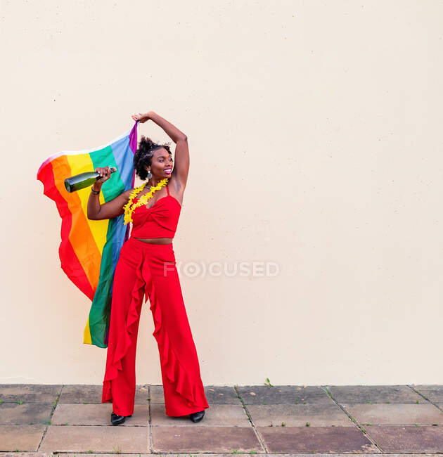 Allegro afroamericano femminile in elegante abbigliamento con bottiglia di bevanda alcolica e bandiera colorata guardando lontano su sfondo chiaro — Foto stock