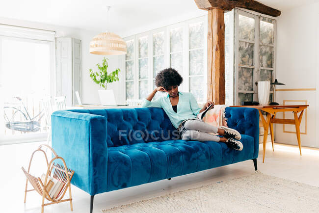 Jovem mulher negra em roupa casual com fones de ouvido sentados no sofá azul acolhedor em casa e revista de leitura enquanto desfruta de tempo livre em casa — Fotografia de Stock