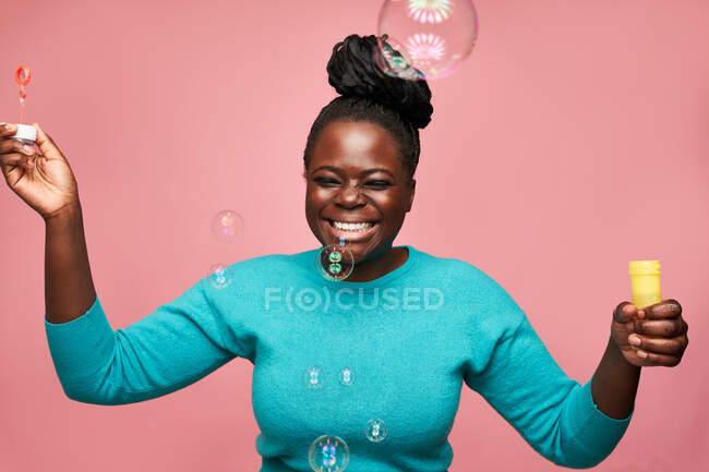 Щаслива афроамериканка з закритими очима в синьому одязі і розмахуючи мильними бульбашками на рожевому тлі в студії — стокове фото