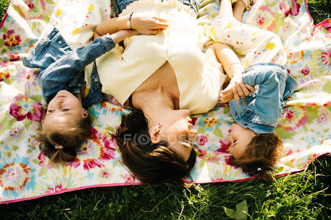 Вид зверху на щасливу молоду жінку і чарівні маленькі сестри в подібних сукнях, лежачи на ковдрі на зеленій траві, проводячи літній день разом у парку — стокове фото