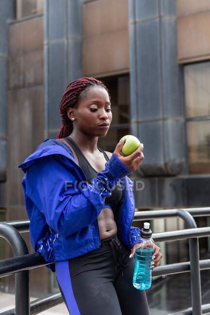 Athletic muscular African-American woman holding apple and sportive drink on street — Stock Photo