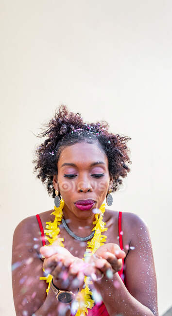 Joven mujer étnica con peinado afro en accesorios que soplan sobre pétalos florales mientras mira hacia abajo sobre fondo claro - foto de stock