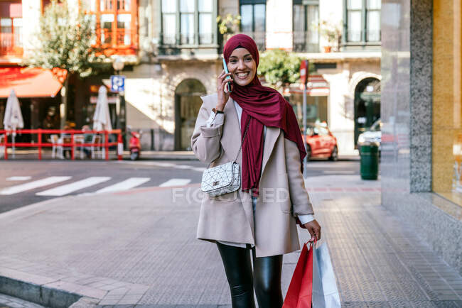 Веселий мусульманин у хіджабі і з торговими сумками, які ходять по вулиці, виступаючи на смартфоні. — стокове фото