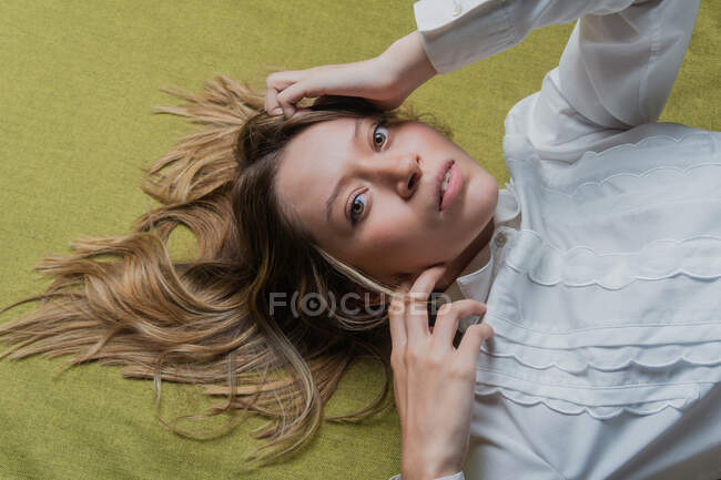 De cima de jovem mulher de olhos verdes com cabelos longos e claros vestindo blusa branca tocando a cabeça e olhando para a câmera enquanto estava deitado em tecido verde — Fotografia de Stock