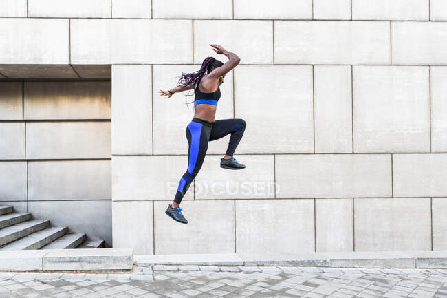 Side view of muscular African American female in sportswear leaping high in air while exercising near wall of modern building on city street — Stock Photo