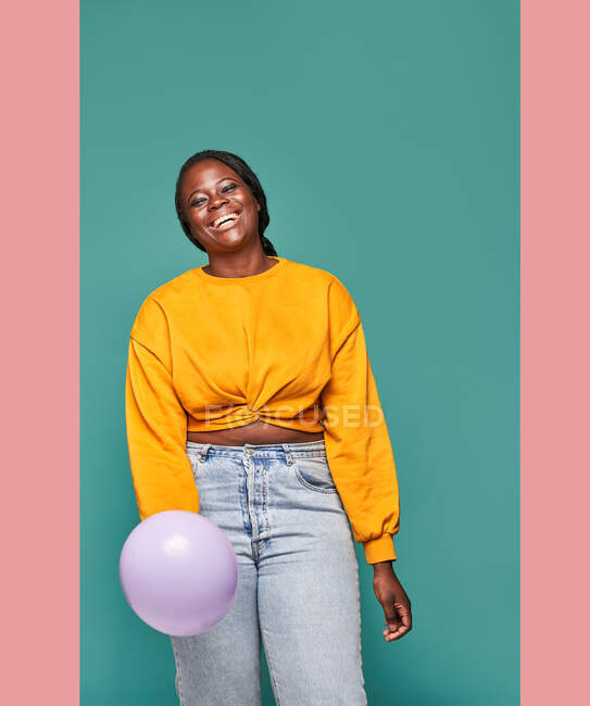 Emocionada mujer afroamericana en jeans y top amarillo de pie cerca de globos cayendo sobre fondo azul en el estudio - foto de stock