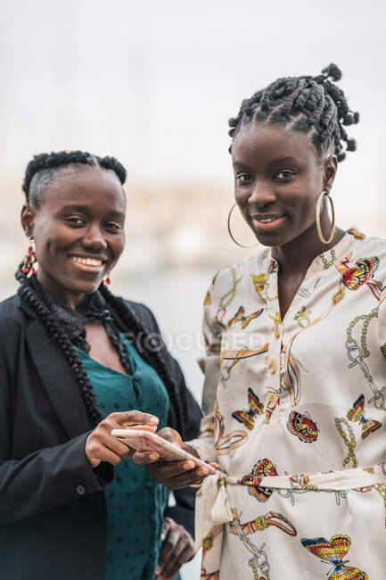 Las damas afroamericanas sonrientes de moda con peinado pasan tiempo juntos navegando por el teléfono móvil en el parque en un día brillante - foto de stock