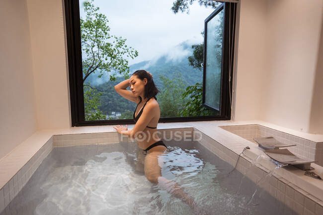 Расслабленная молодая этническая леди в купальниках, сидящая в японской ванне в спа-курорте рядом с окном с видом на горы и зеленые деревья — стоковое фото