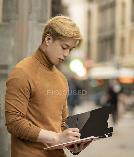 Focused ethnic male entrepreneur standing in street and writing on paper n folder — Stock Photo