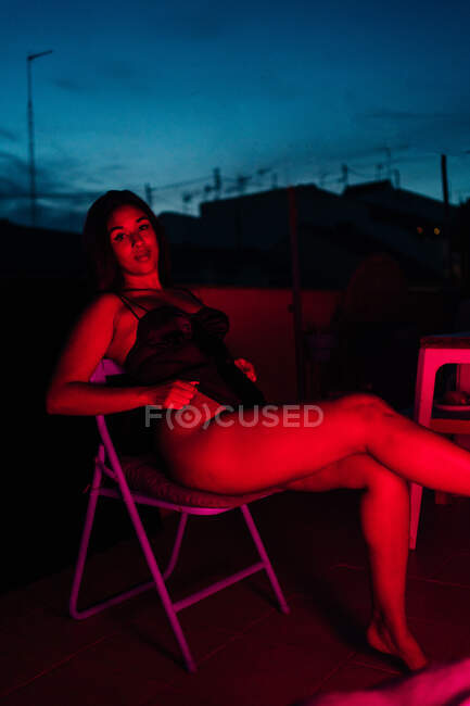 Jeune femme en sous-vêtements regardant la caméra assis sur une chaise sous la lumière rouge néon la nuit sur la terrasse — Photo de stock