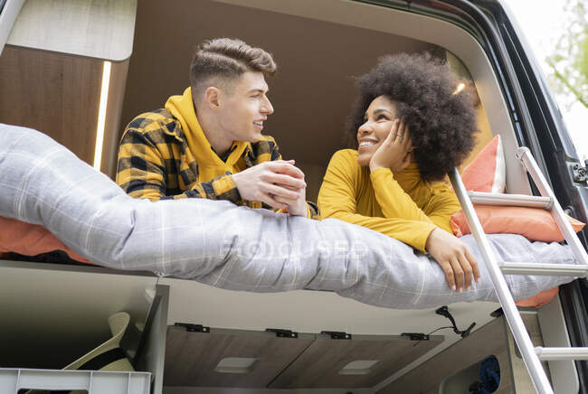 Bajo ángulo de la mujer afroamericana sonriendo para el novio joven mientras se acuesta en la cama en la furgoneta durante el viaje por carretera mirándose - foto de stock