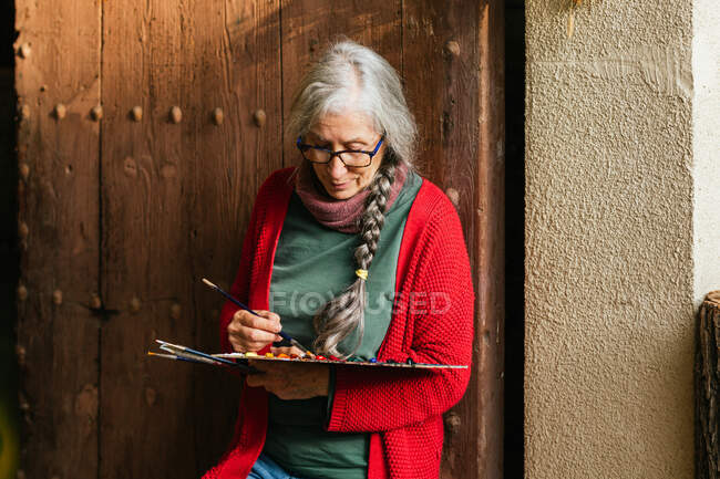 Encantada artista feminina sênior com paleta de tinta e pincéis de pé perto da porta de madeira — Fotografia de Stock