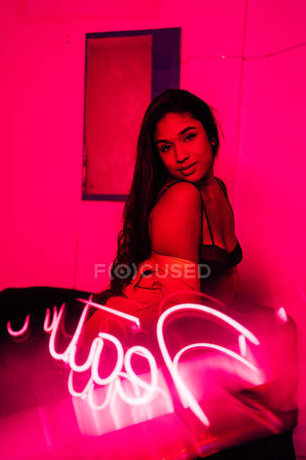 Young Hispanic female touching hair and looking at camera while leaning on wall near neon inscription — Stock Photo