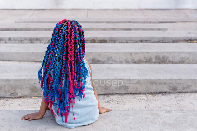 Vista posteriore di irriconoscibile bambino etnico sognante con trecce colorate sedute su scale di cemento mentre guarda lontano alla luce del giorno — Foto stock
