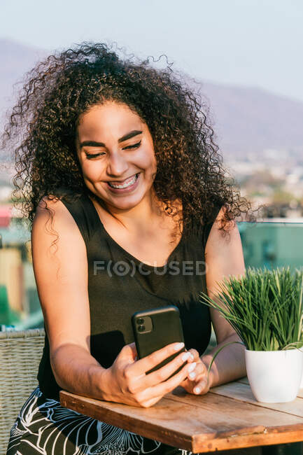 Fröhliche junge lockige hispanische Frau plaudert auf dem Handy, während sie sich an einem sonnigen Sommerabend auf der Caféterrasse ausruht — Stockfoto
