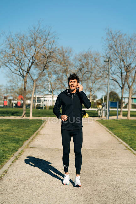 Взрослый бегун в спортивной одежде бегает по тротуару между газонами и смотрит вперед во время тренировки в городе — стоковое фото