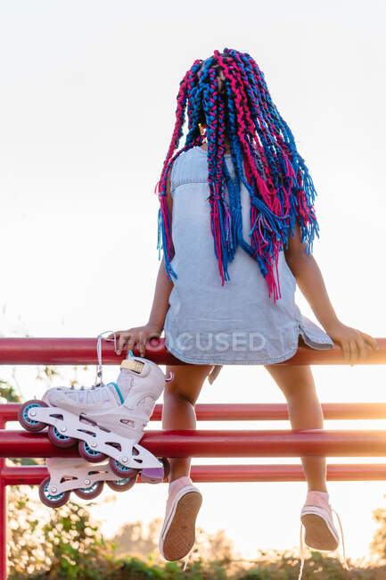 Back view of anonymous Mexican kid with colorful braids admiring trees while leaning on fence in evening — Stock Photo