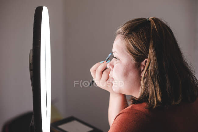 Вид сбоку пухлой женщины с помощью кисти для нанесения макияжа рядом с кольцевым светом в студии — стоковое фото