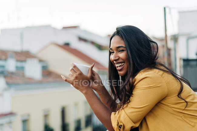 Side view of young cheerful Hispanic woman with closed eyes enjoying hot beverage while resting on balcony in evening — Stock Photo