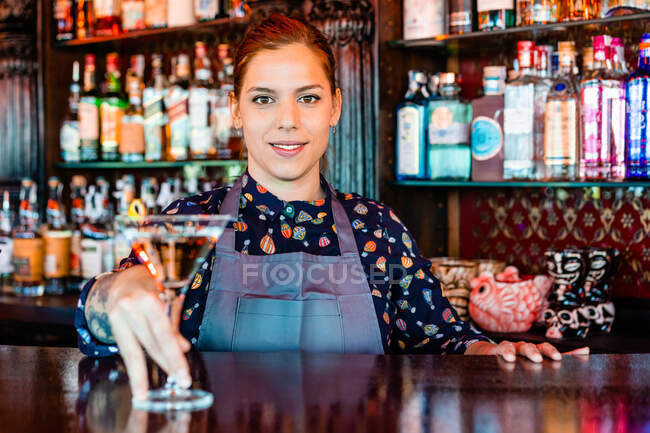 Cheerful female barkeeper with glass of refreshing vermouth standing at counter in bar and looking at camera — Stock Photo