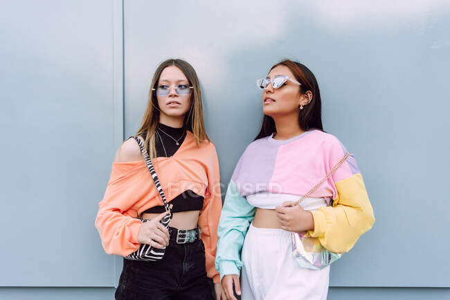 Namoradas confiantes vestindo óculos de sol na moda e roupas casuais modernas de pé juntos perto da parede e olhando para longe — Fotografia de Stock