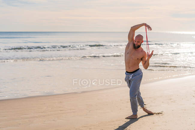 Side view of shirtless male athlete stretching arms with elastic band while working out on empty sunny beach — Stock Photo