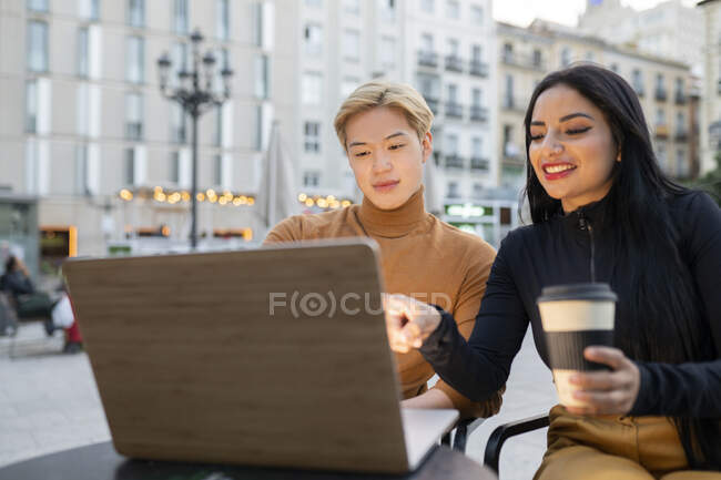 Fröhliche multiethnische Freiberuflerin sitzt mit Laptop am Tisch und arbeitet gemeinsam an einem Remote-Projekt im Café im Freien — Stockfoto
