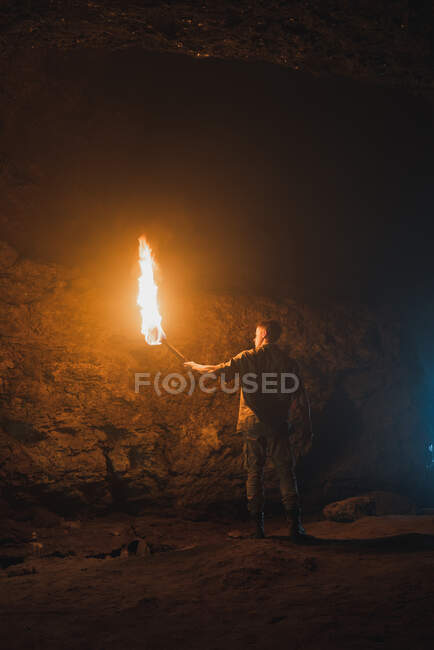 Vista posterior del joven espeleólogo masculino con antorcha encendida de pie en la oscura cueva rocosa estrecha mientras explora el entorno subterráneo - foto de stock