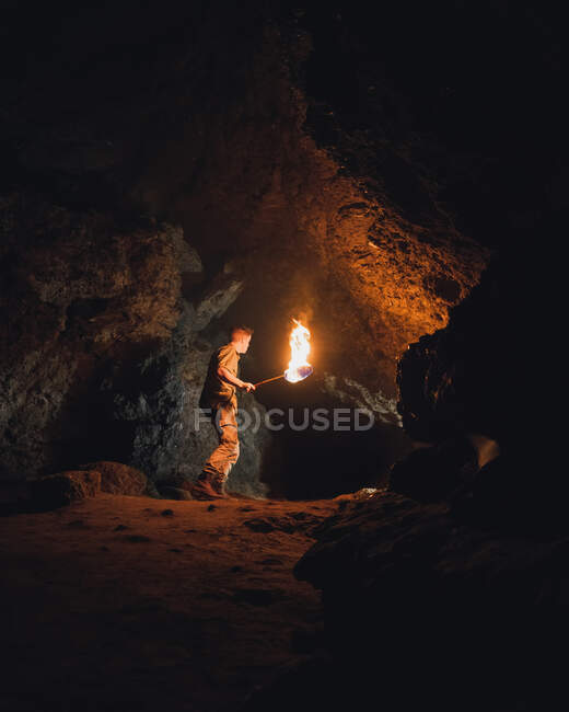 Side view of young male speleologist with flaming torch standing in dark narrow rocky cave while exploring subterranean environment — Stock Photo