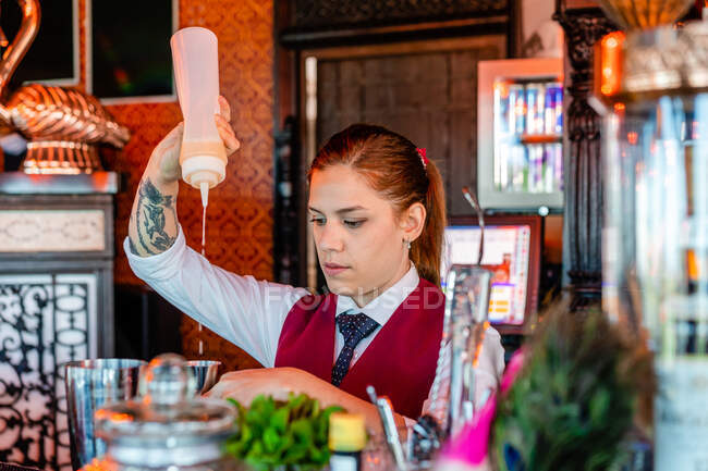 Barman feminino grave em uniforme de pé no balcão no bar e derramando licor em agitador enquanto prepara coquetel de álcool — Fotografia de Stock