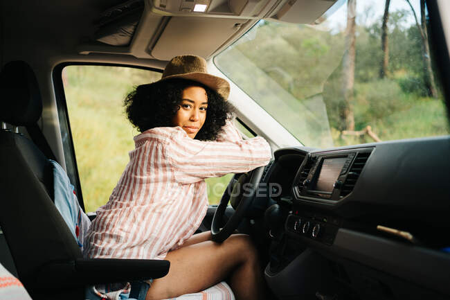Vista lateral do romântico pensativo jovem afro-americano viajante feminino sentado no assento do motorista e olhando para a câmera enquanto desfruta de viagem de verão na natureza — Fotografia de Stock
