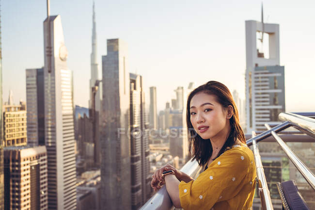 Giovane donna asiatica viaggiatore sulla terrazza panoramica guardando la fotocamera sorridente contro vista mozzafiato della città di Dubai con l'architettura contemporanea — Foto stock