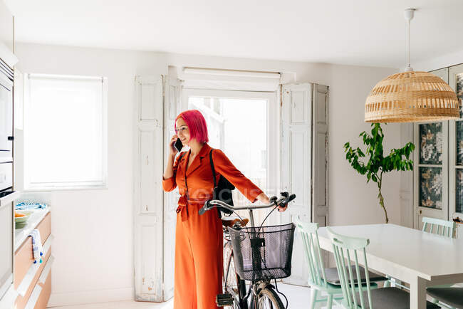 Giovane designer creativa donna in abito alla moda e occhiali parlare su smartphone mentre in piedi con la bicicletta in un moderno appartamento leggero — Foto stock