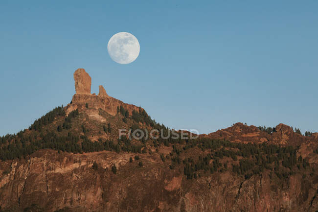 Paisaje espectacular con gran luna llena en el cielo azul sobre el pico rocoso de la montaña con bosque verde en la noche de verano - foto de stock