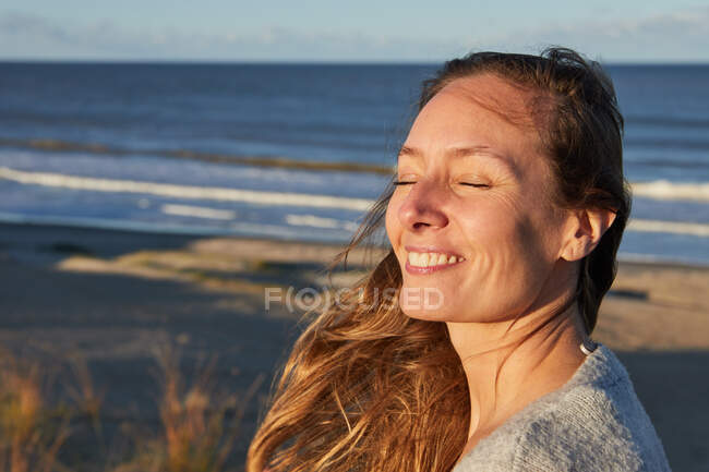 Mujer tranquila con los ojos cerrados disfrutando de la puesta de sol en verano en la playa cerca del mar - foto de stock