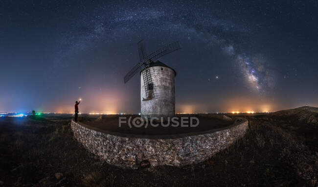 Cenário pitoresco da Via Láctea no céu noturno escuro acima da torre de moinho de vento de pedra envelhecida com luzes brilhantes à distância — Fotografia de Stock