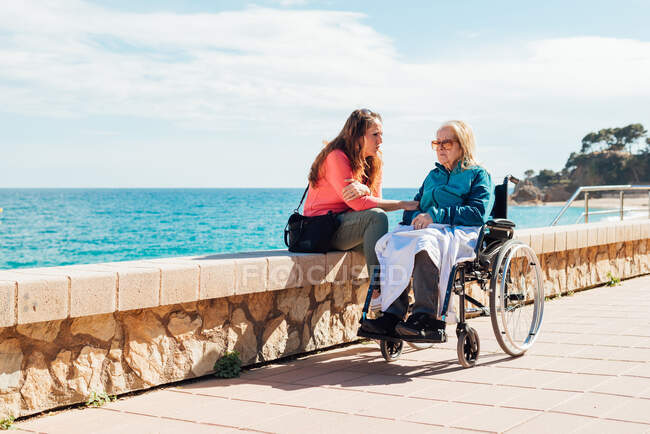 Mujer adulta y madre anciana en silla de ruedas enfriándose en el terraplén y hablando entre sí en un día soleado cerca del mar - foto de stock