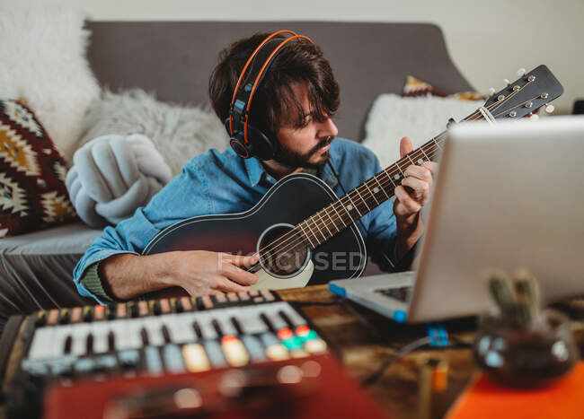 Hombre joven en auriculares tocando en la guitarra cerca de la mesa con el ordenador portátil y sintetizador en casa mirando a la cámara - foto de stock