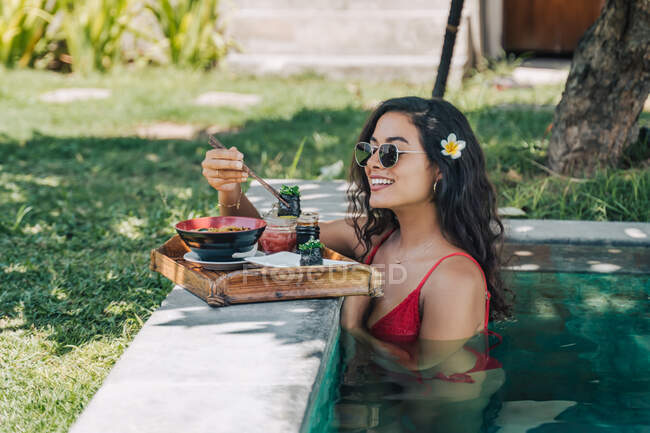 Vista lateral de la alegre viajera femenina en traje de baño contra la piscina con deliciosa pasta asiática cocinada entre palillos a la luz del sol - foto de stock