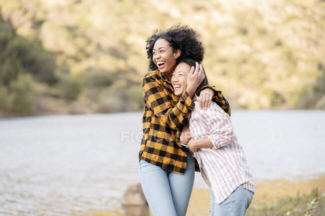 Conteúdo LBGT casal de mulheres multirraciais abraçando-se enquanto caminham com o cão ao longo do lago na floresta olhando para longe — Fotografia de Stock