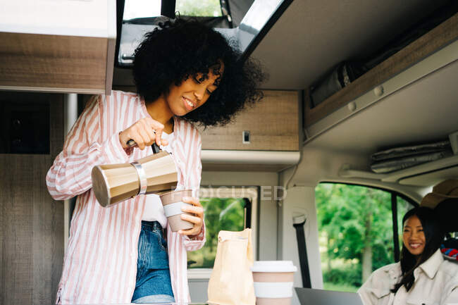 Positive junge Afroamerikanerin gießt Kaffee aus Wasserkocher in Tasse, während sie mit asiatischer Freundin in Wohnmobil während Sommerreise in die Natur rastet — Stockfoto
