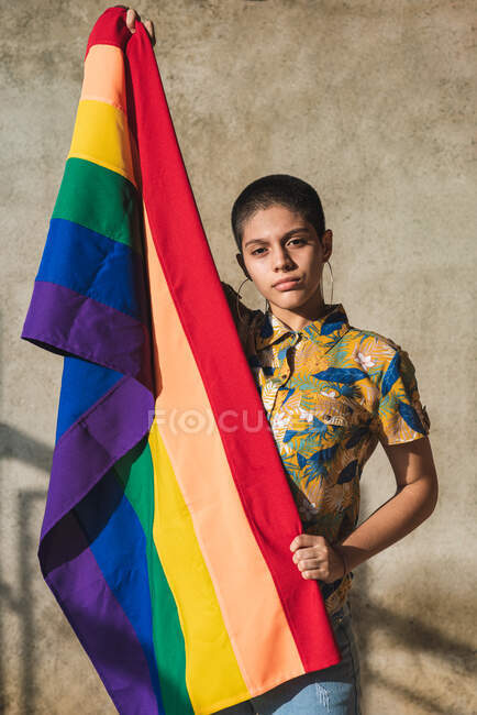 Mujer étnica bisexual joven seria con bandera multicolor que representa símbolos LGBTQ y mira a la cámara en un día soleado - foto de stock